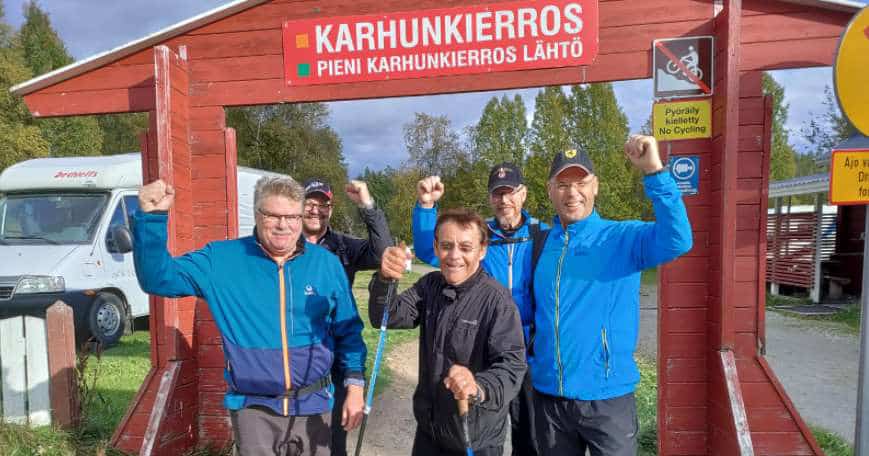 Viisi iloista miestä hymyilee kädet ylhäällä, takana on punainen kyltti, jossa lukee Karhunkierros