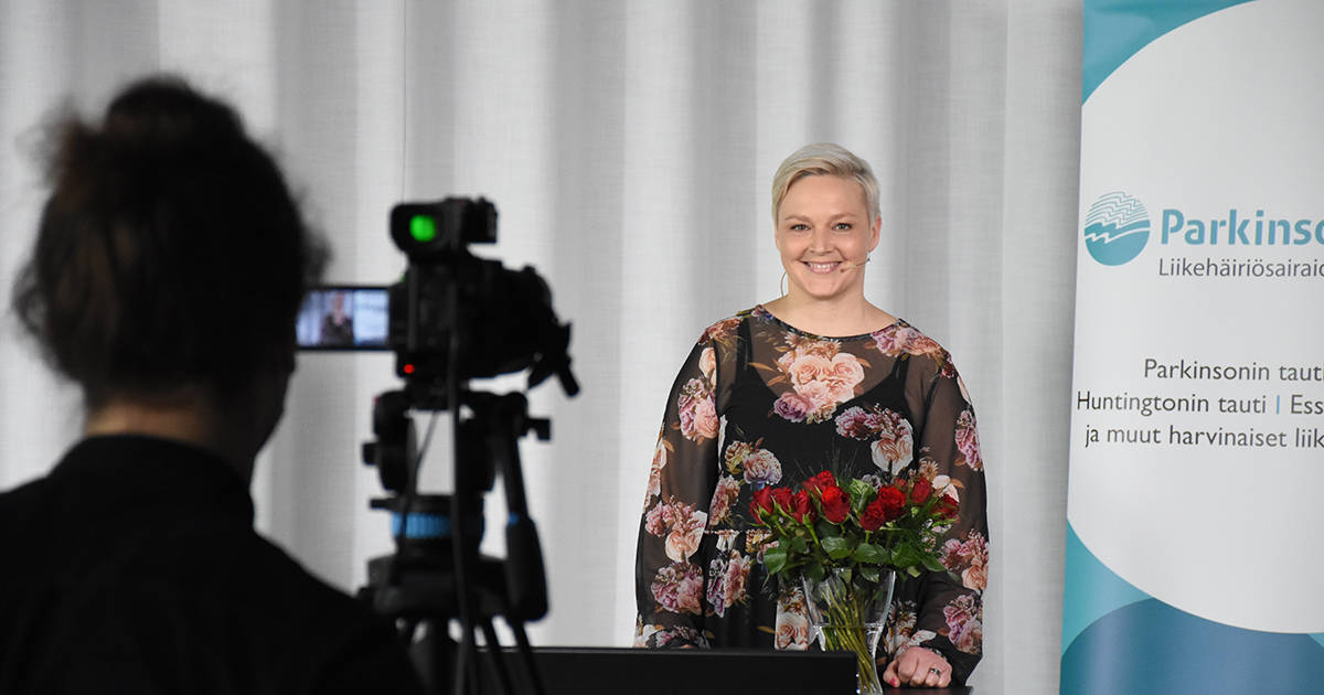 Vaaleahiuksinen nainen seisoo ja hymyilee kameraan, kuvan etualalla näkyy kamera jalustalla