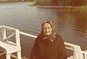 Vanha huivipäinen nainen istuu laiturilla, taustalla järvi ja kevään alkava kaislikko