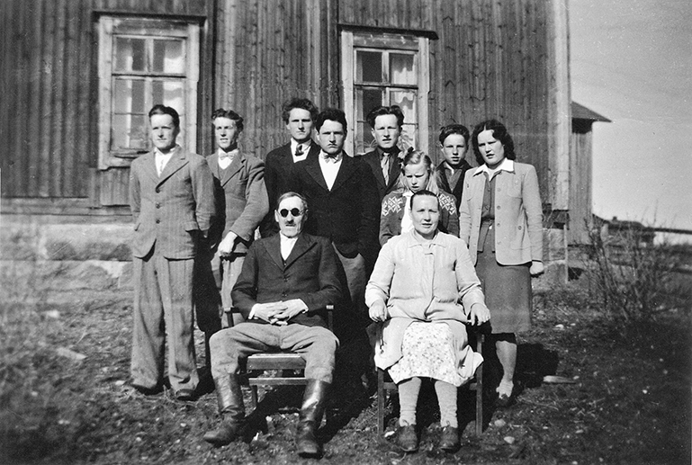 Vanhassa mustavalkokuvassa kuusi miestä ja nainen seisovat vanhan talon edessä, heidän edessään istuvat tuoleilla vanha isäntä ja emäntä ja katsovat kameraan.