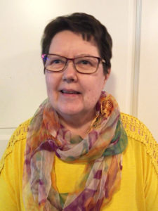 Keski-ikäinen nainen, jolla on keltainen paita, värikäs huivi, silmälasit ja tummat lyhyet hiukset katsoo hieman ohi kameran ja hymyilee.